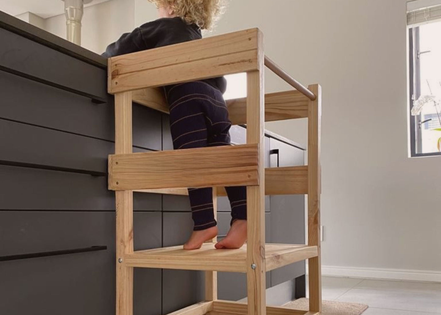 Toddler standing safely in a handcrafted wooden Learning Tower at a kitchen counter, encouraging independence and family connection