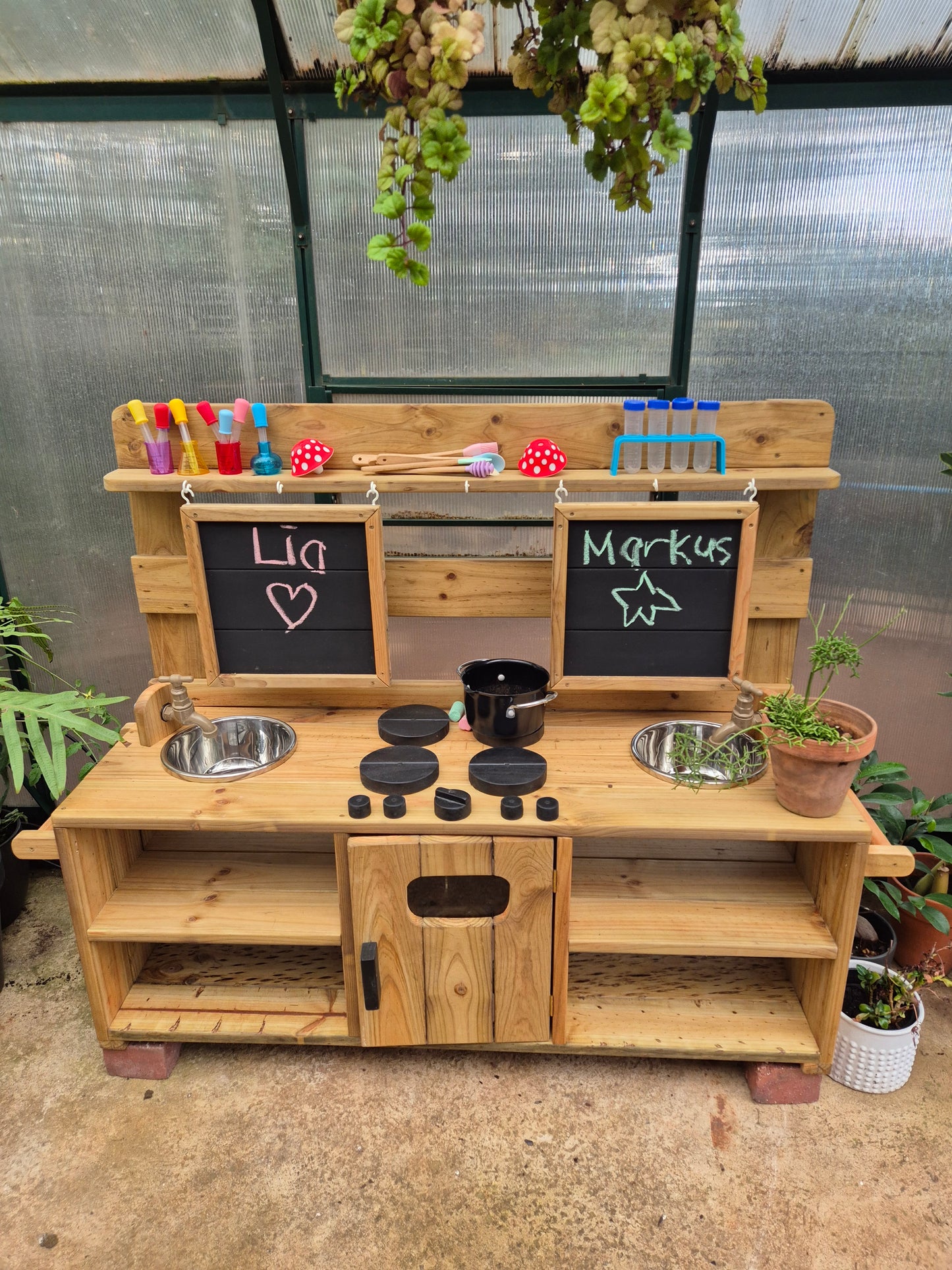 Double wooden mud kitchen with two sinks, working taps, chalkboards, hob and storage, set up for children’s outdoor play in a greenhouse.