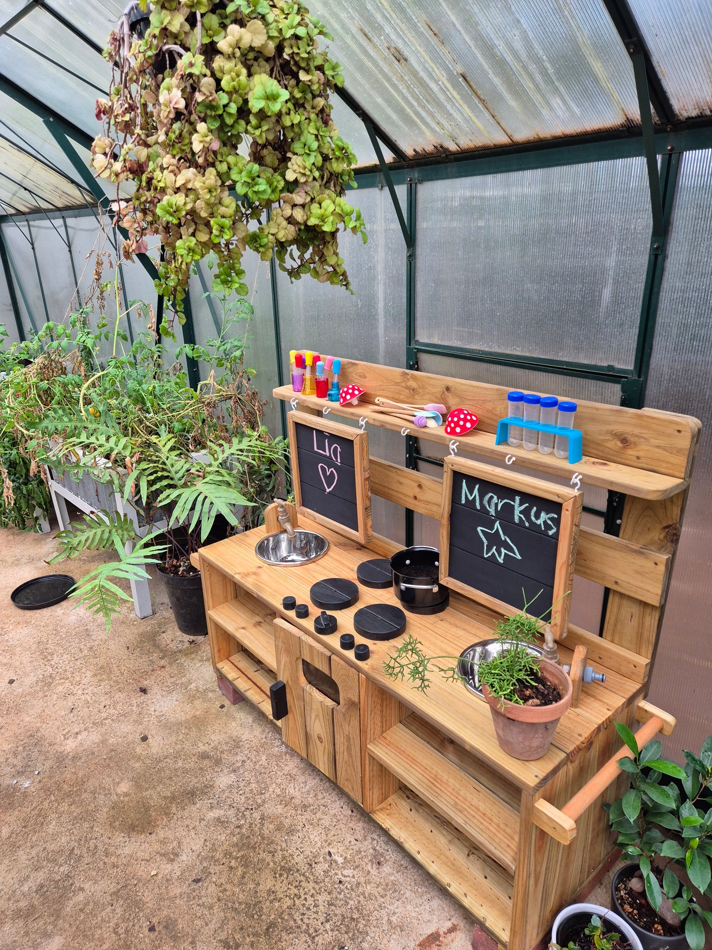 Double wooden outdoor mud kitchen with two sinks, taps, chalkboards, hob, oven and open storage, set up with play accessories and plants in a greenhouse.