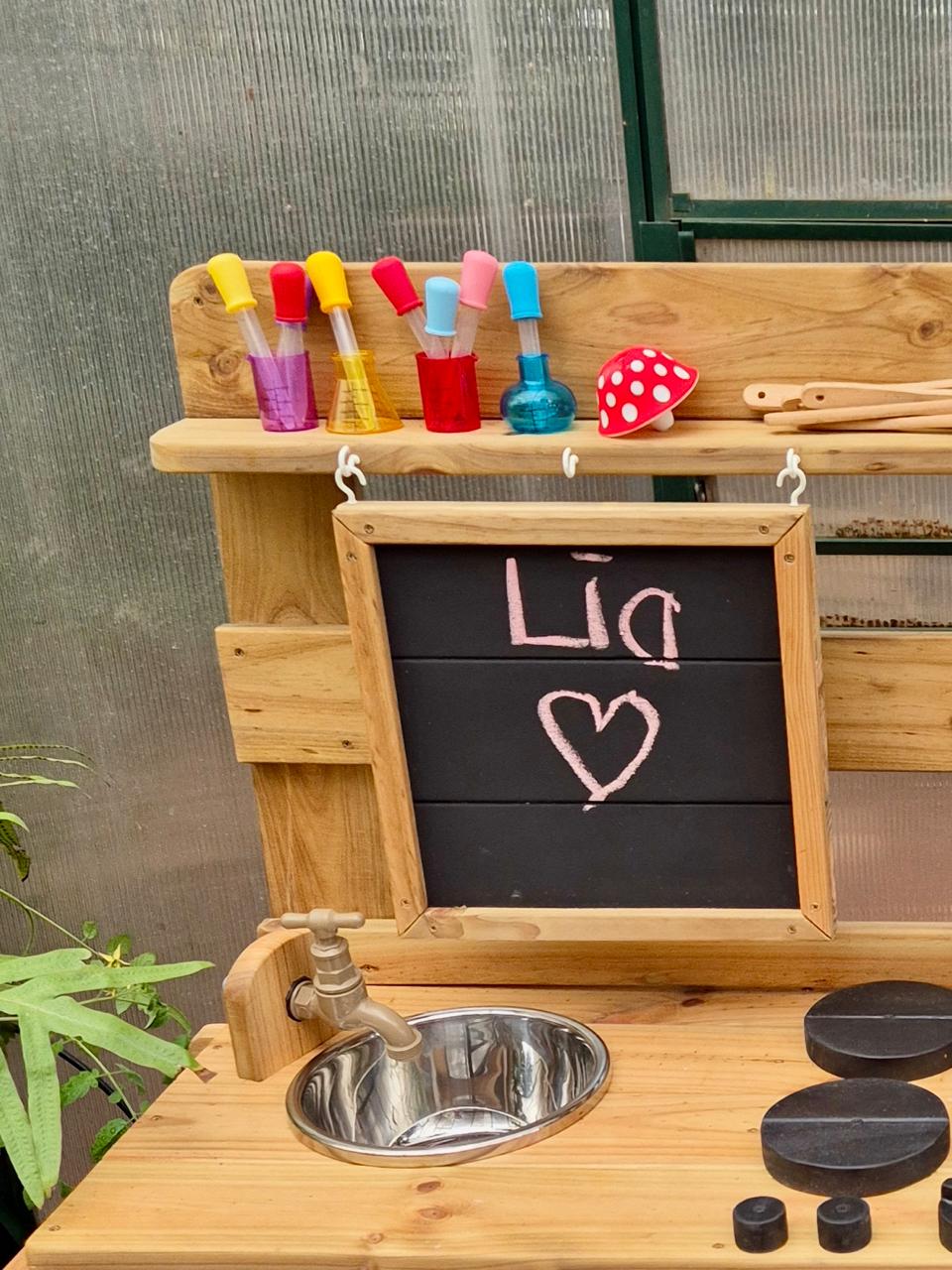 Close-up of a handcrafted wooden Single Mud Kitchen with working tap, stainless steel bowl, chalkboard, play hobs and colourful pretend potion bottles.