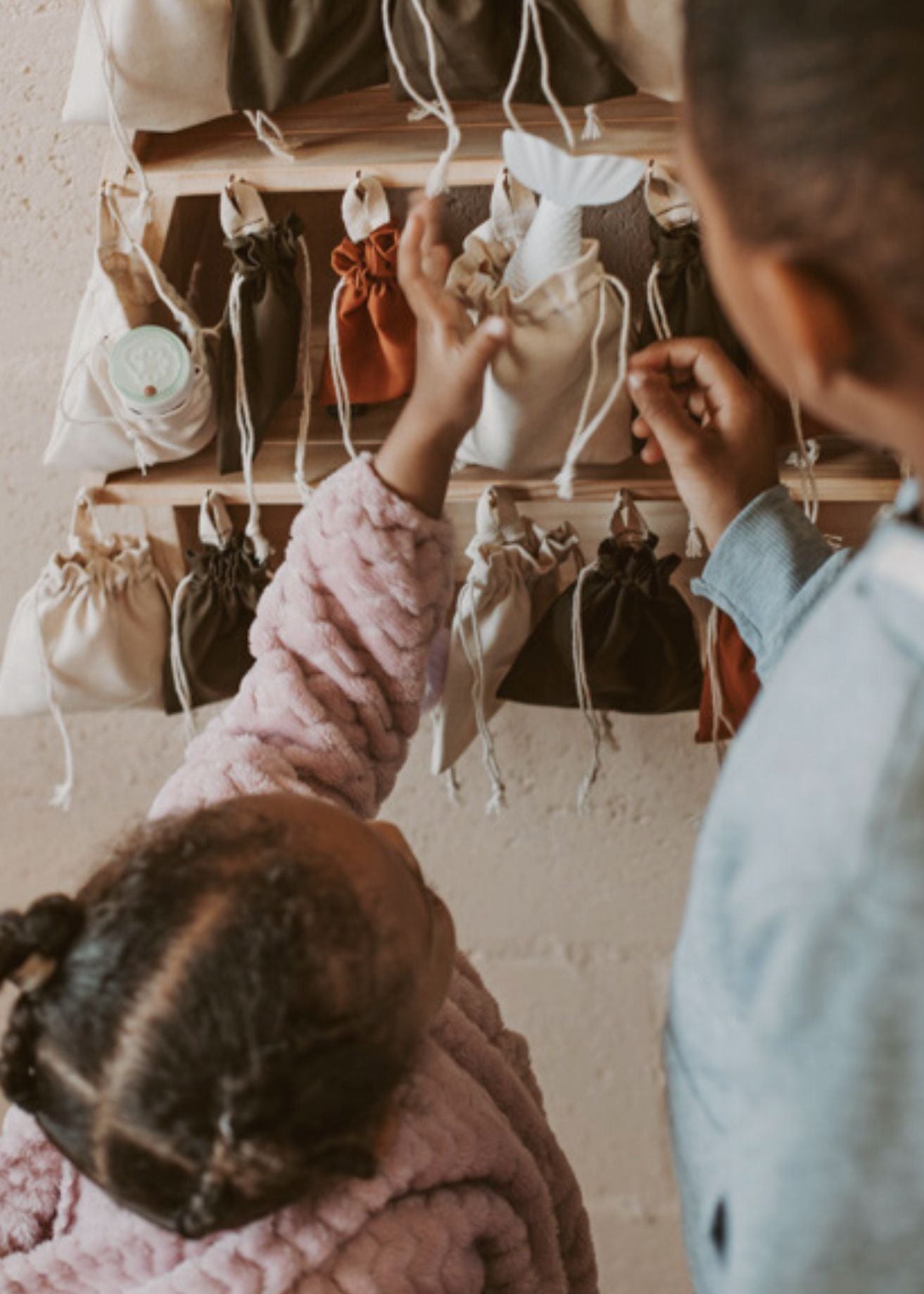 Two young children reaching into fabric drawstring bags on a wooden advent calendar, choosing surprises for the Christmas countdown