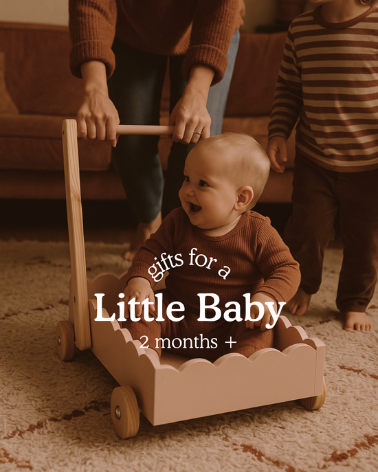 Baby sitting inside a wooden scalloped walker toy, smiling as an adult supports the handle behind them. Text on image reads ‘Gifts for a Little Baby, 2 months +