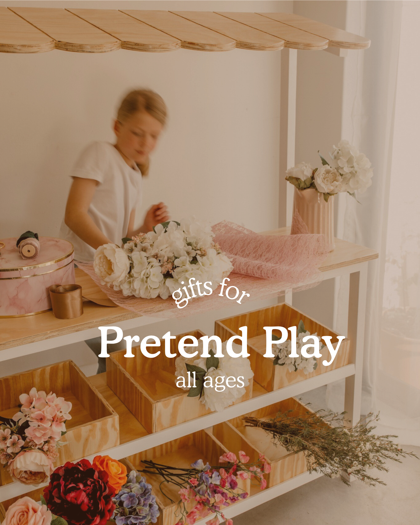 Child playing at a wooden market stall filled with pretend flowers and accessories in a bright, airy room. Text on image reads ‘Gifts for Pretend Play, all ages
