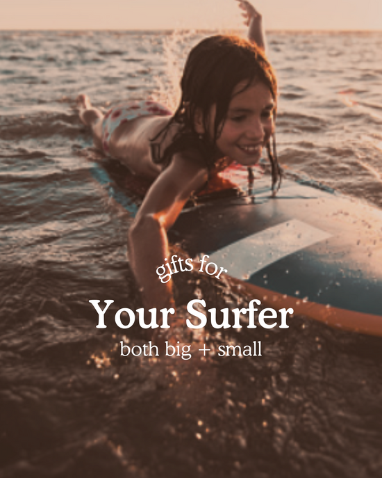 Child lying on a surfboard and paddling through the ocean at sunset, smiling as they ride the water. Text on image reads ‘Gifts for Your Surfer, both big + small'