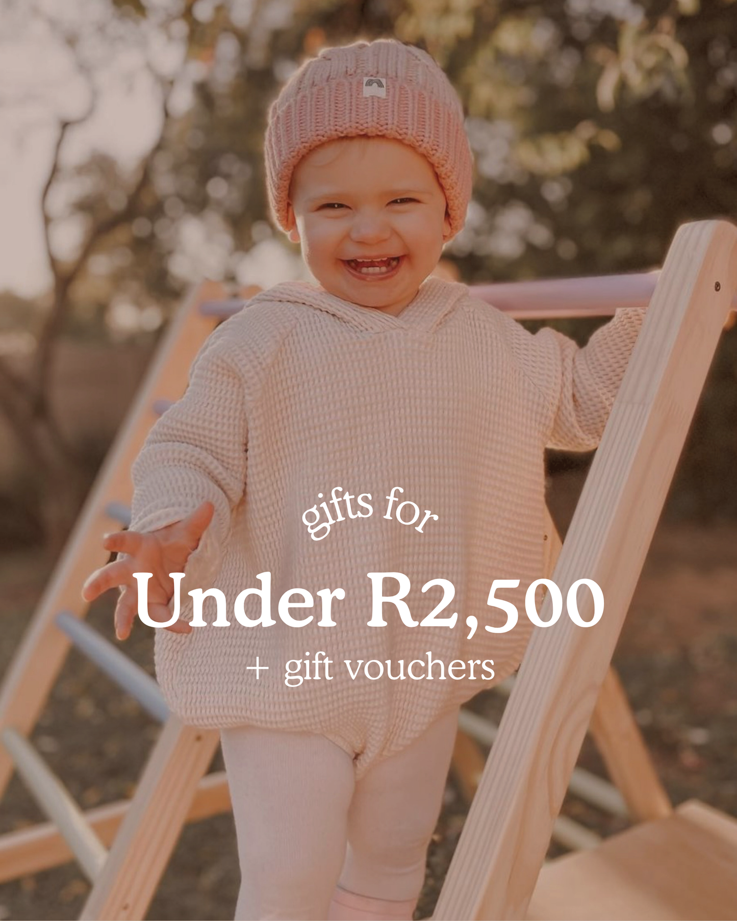 Smiling toddler standing outdoors on a wooden climbing frame, wearing a pink beanie and cosy knit outfit. Text on image reads ‘Gifts for Under R2,500 + gift vouchers