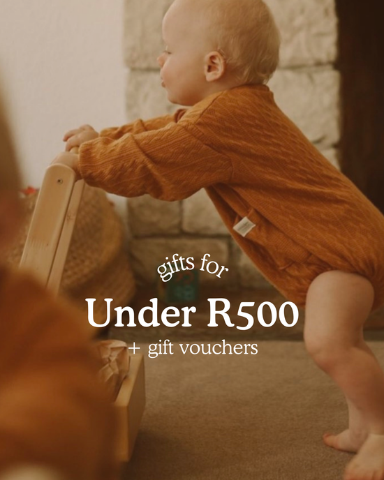 Baby standing and holding onto a wooden toy in a cosy room. Text on image reads ‘Gifts for Under R500 + gift vouchers’