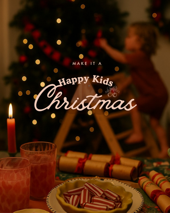 Festive Christmas scene with candles, crackers, sweet treats and ornaments on a holiday table, with a child decorating a Christmas tree in the background. Overlay text reads ‘Make it a Happy Kids Christmas’