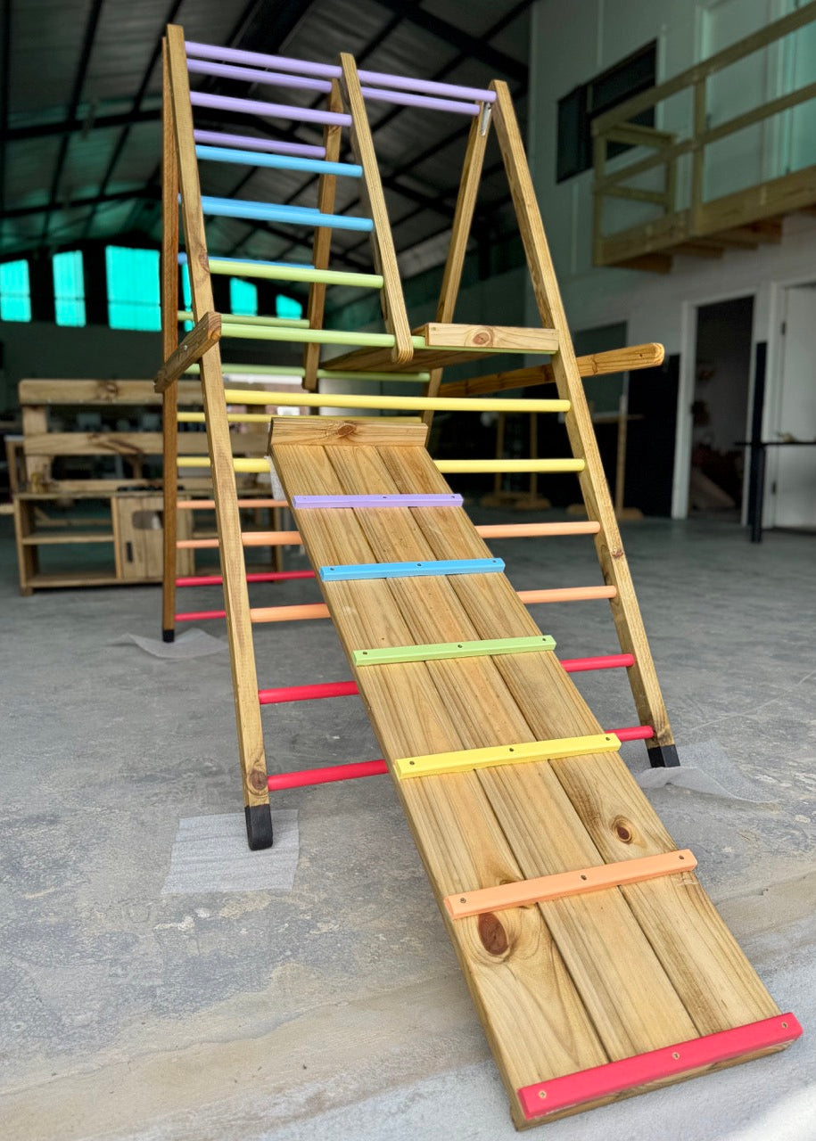 Outdoor Pikler Playground with rainbow-coloured climbing rungs and a wooden slide, shown from a front-angle view in the workshop