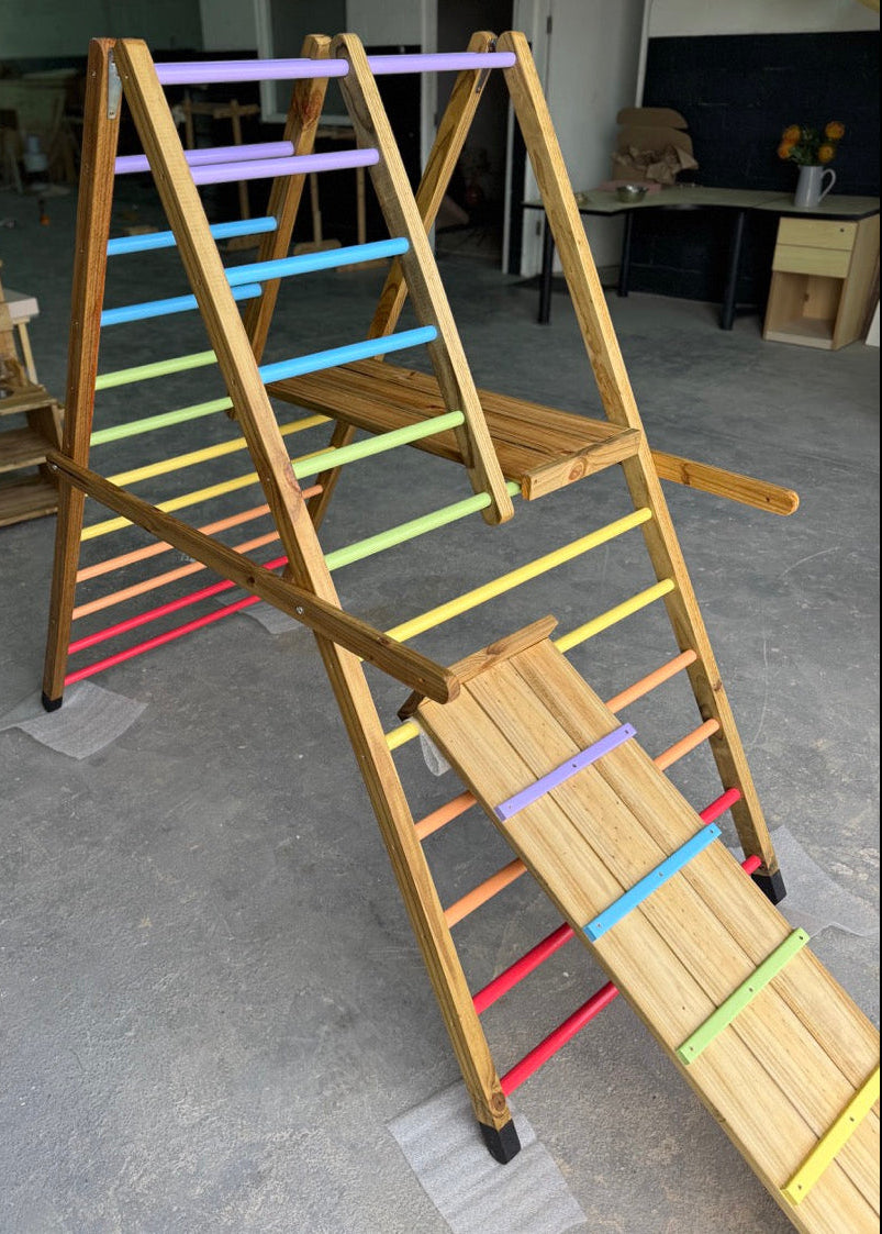 Angled view of the Outdoor Pikler Playground with the reversible ramp in the climbing-ramp position, featuring pastel-coloured grip rungs and rainbow ladder bars on a treated-timber frame.
