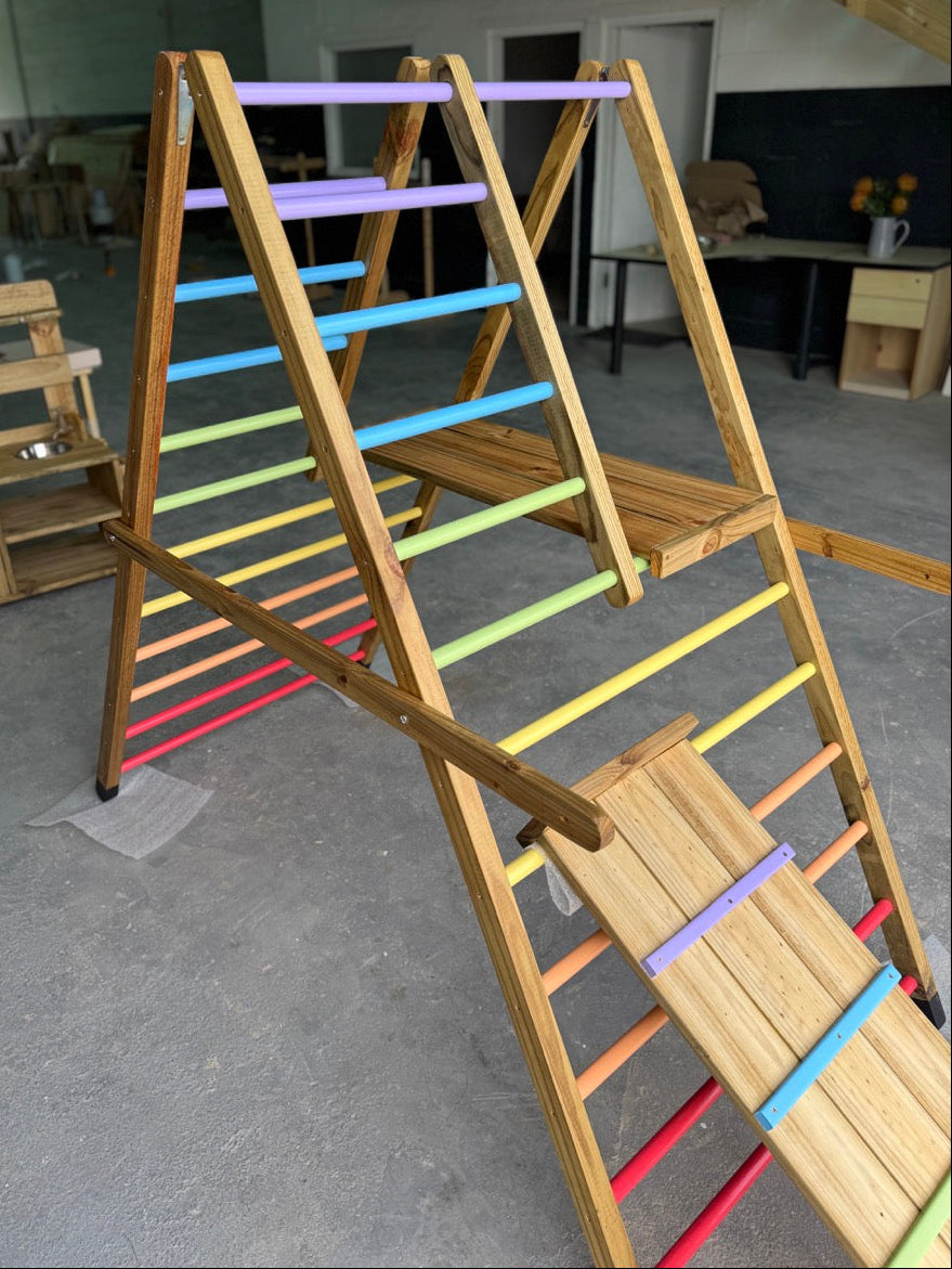 High-angle view of the Outdoor Pikler Playground with the reversible ramp in the climbing-ramp position, showing pastel-coloured grip rungs and rainbow ladder bars on the treated-timber frame.