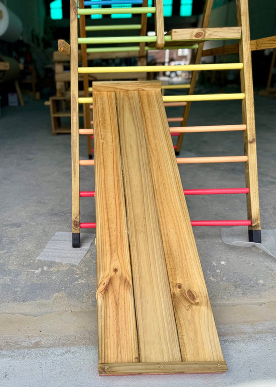 Close-up of the smooth slide side of the reversible ramp on the Outdoor Pikler Playground, showing the treated timber surface and rainbow-coloured rungs.