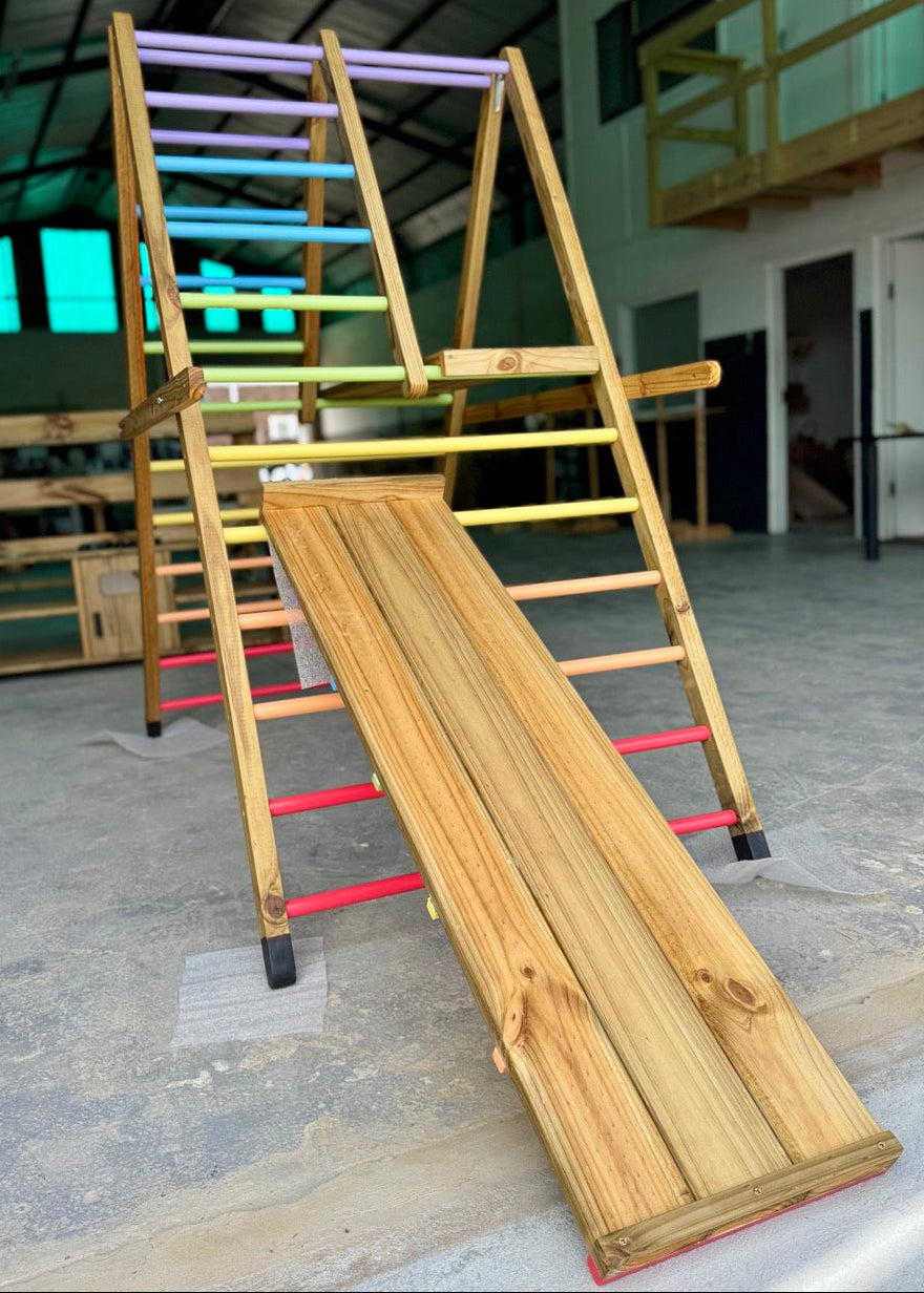 Front-angle view of the Outdoor Pikler Playground with the reversible ramp in slide mode, showing the smooth timber slide surface and pastel rainbow climbing rungs