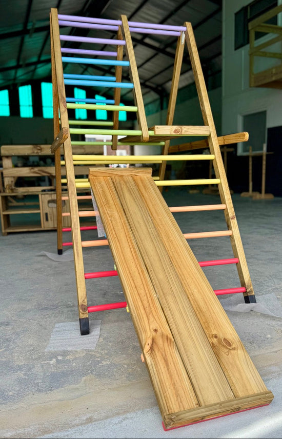 Front view of the Outdoor Pikler Playground with the reversible ramp in slide mode, showing the smooth timber slide surface and pastel rainbow climbing rungs on the treated-timber frame.