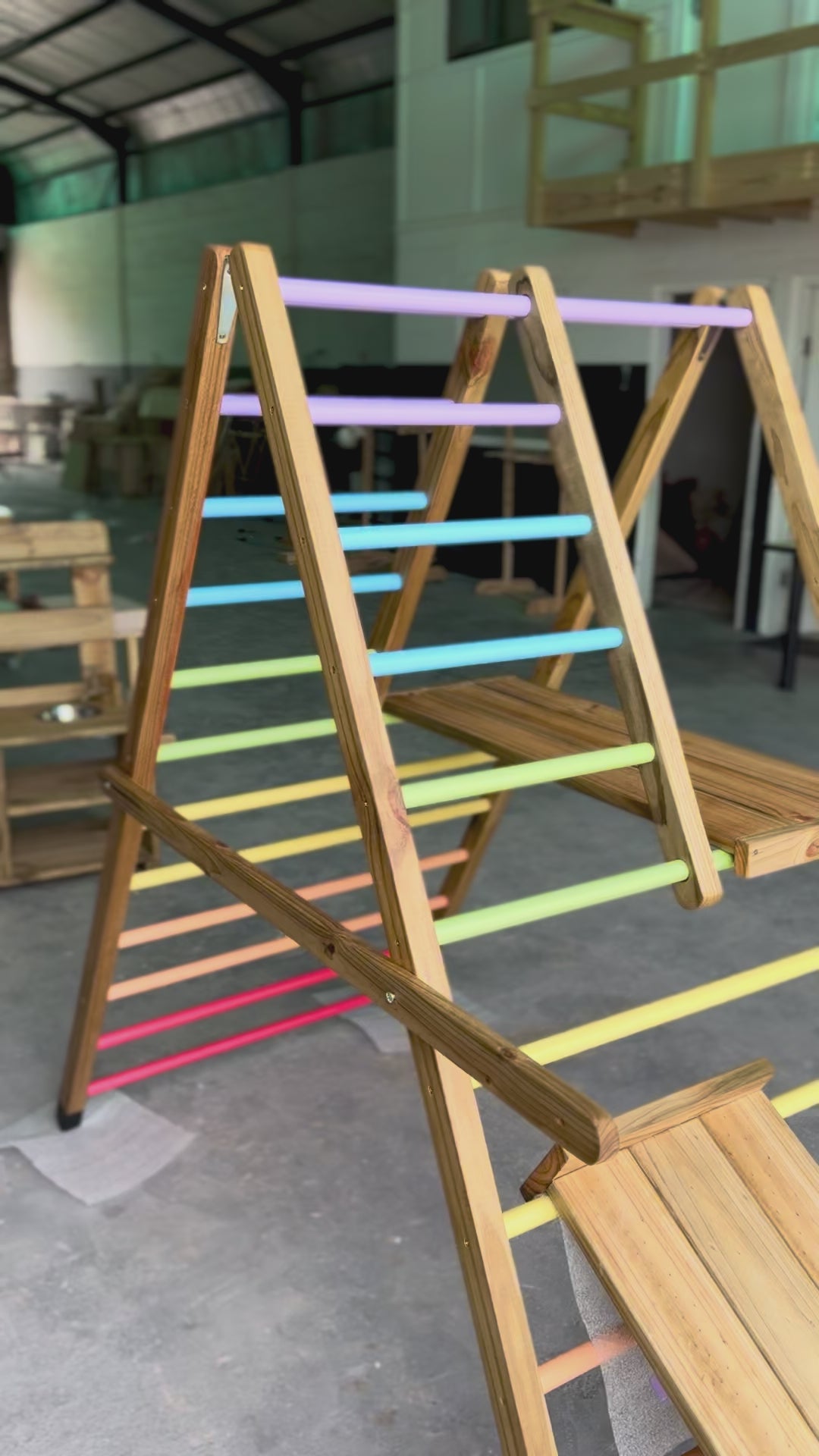 Load and play video in Gallery viewer, Video starting with a tight side view of the Outdoor Pikler Playground, then stepping back to reveal the full reversible ramp in slide mode, with rainbow-coloured climbing rungs and a treated-timber frame.
