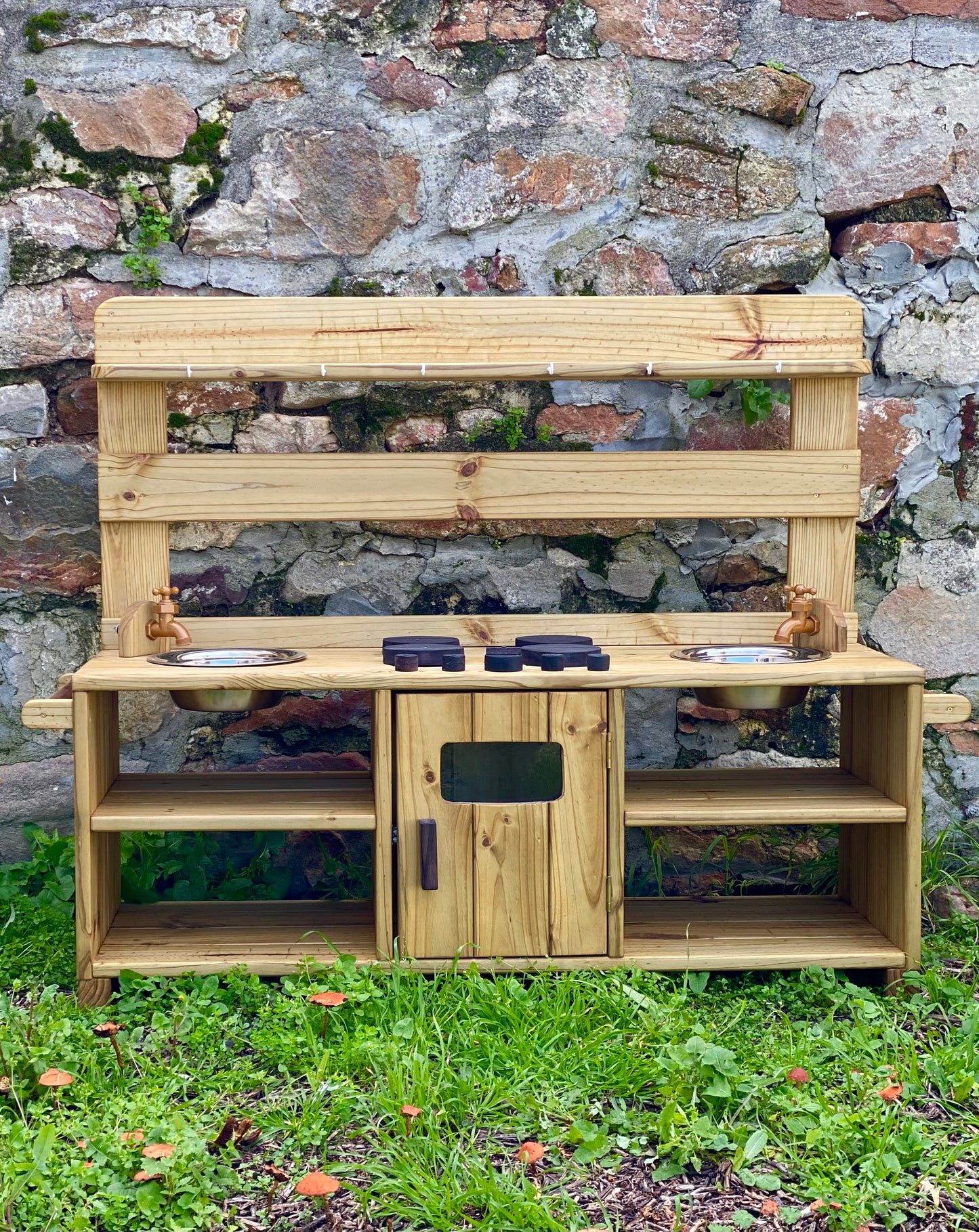 Front view of the handcrafted Double Mud Kitchen against a rustic stone wall, featuring twin taps, sinks, and wooden shelves.